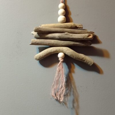 Suspension murale en bois flotté avec perles blanches et pompon rose pastel sur fond gris.