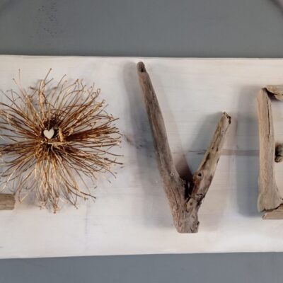 Pancarte en bois avec le mot "LOVE" formé de bois flotté et d'une fleur sèche, suspendue par une corde de jute.
